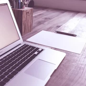 A desk with a laptop, a piece of paper, a pen and a pen holding box in the background